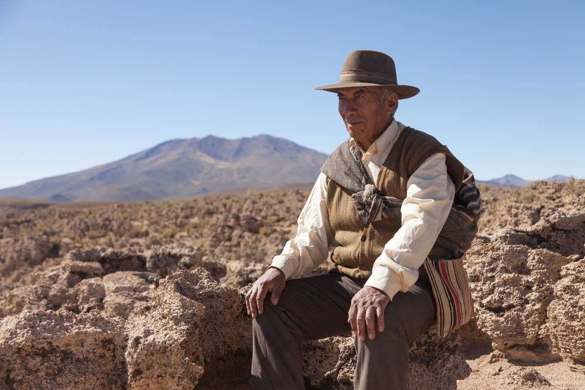 Ein Mann mit einem Hut sitzt auf einem Stein in einer Wüste. Im Hintergrund ist ein Berg zu sehen.