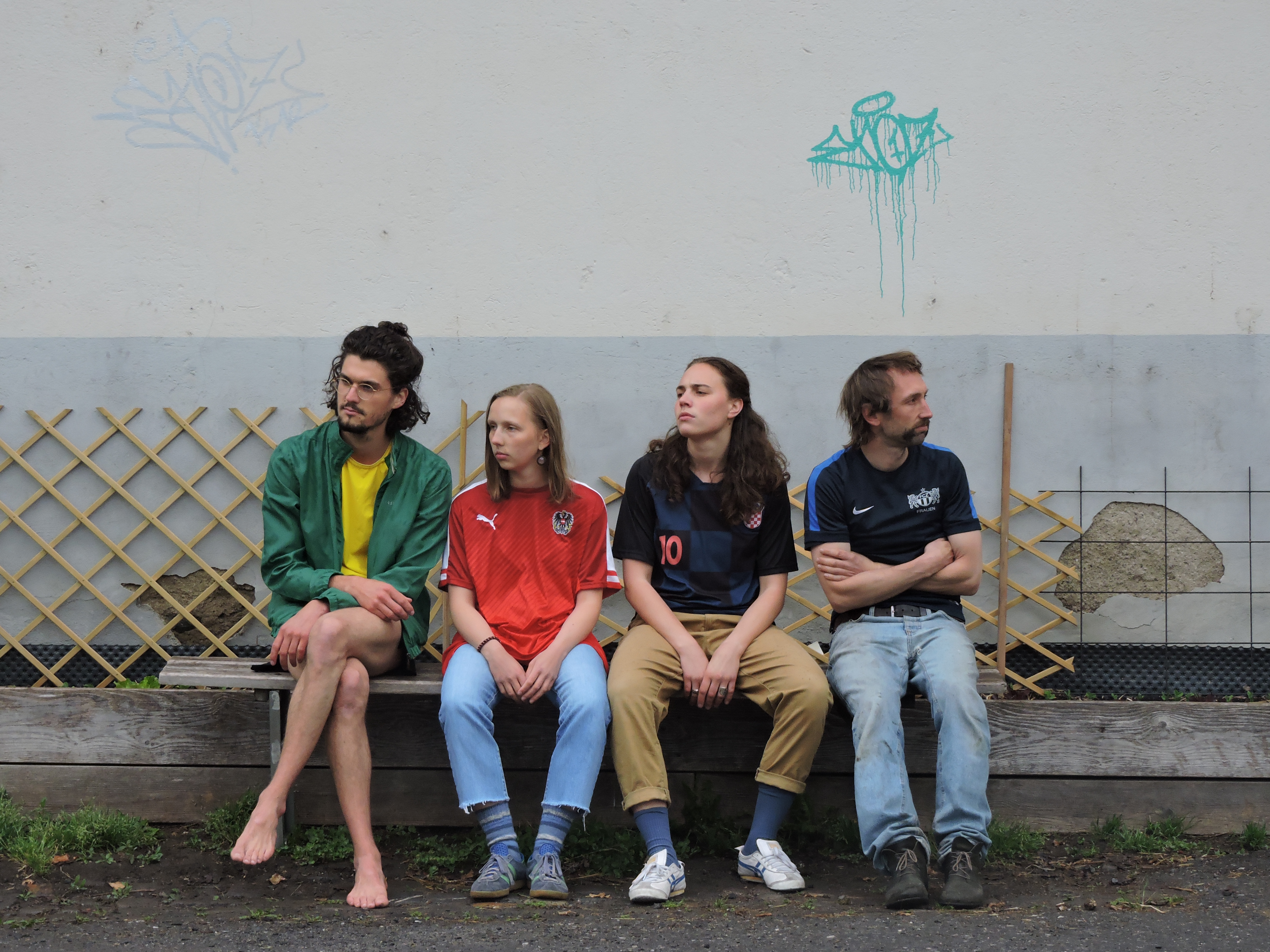 Vier Menschen sitzen auf einer Bank und schauen in verschiedene Richtungen.. Sie tragen alle Fussballshirts. Es ist ein Bandfoto.
