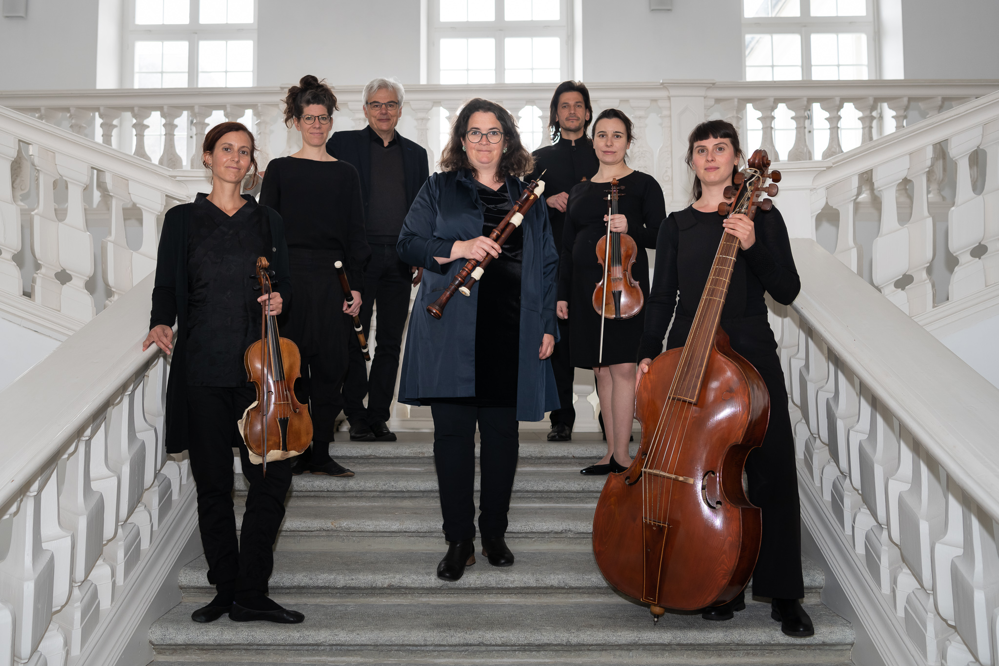 Fünf Frauen und zwei Männer stehen auf einer Treppe mit ihren Instrumenten in der Hand und schauen leich nach unten in die Kamera.