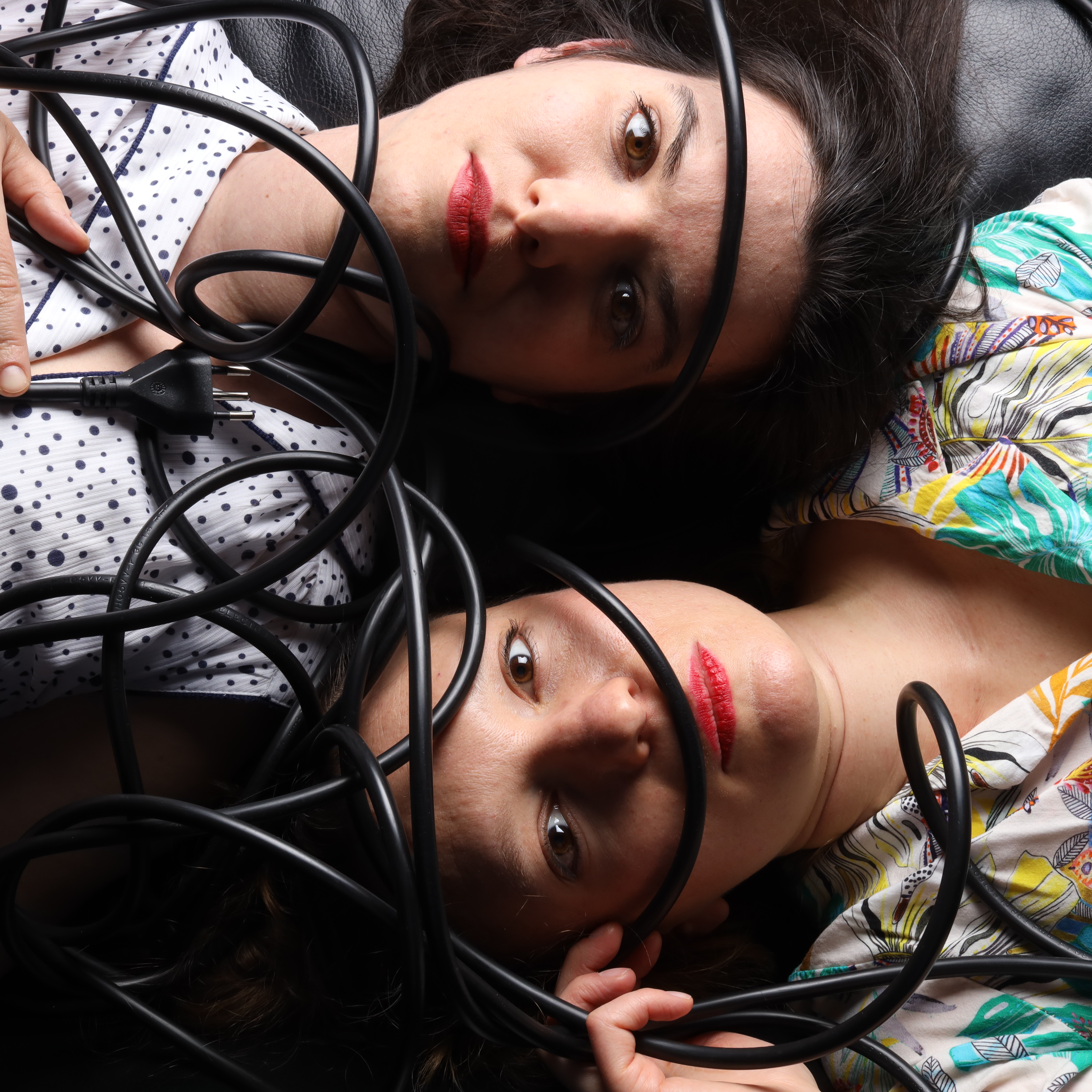 Zwei Frauen liegen kopf-an-Kopf und schauen in die Kamera. Über ihnen liegt ein schwarzes Stromkabel.