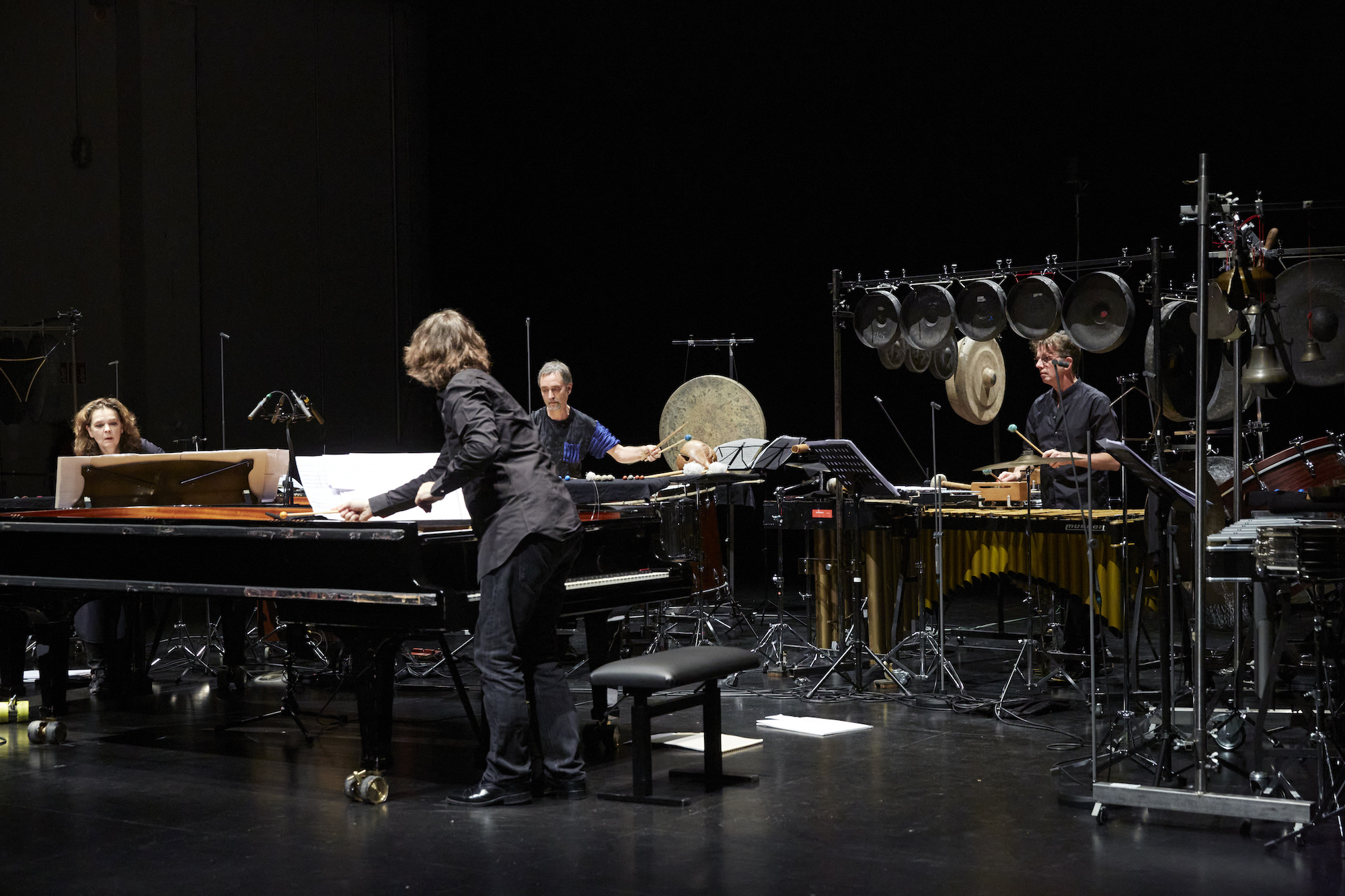 Diverse Instrumente (Klavier, Schlaginstrumente, ect.) stehen auf dunkler Bühne. Vier Personen, zwei Männer und zwei Frauen, spielen auf den Instrumenten. Das Bild ist sehr dunkel.