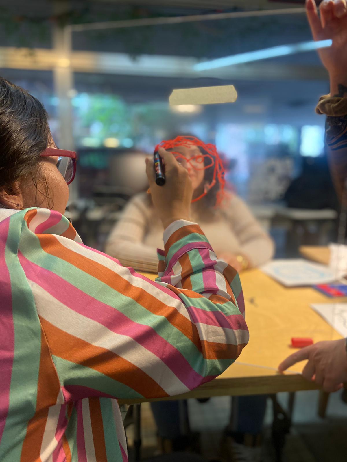 Es sind zwei Personen sichtbar. Eine Person zeichnet die andere ab auf einem Plexiglas.
