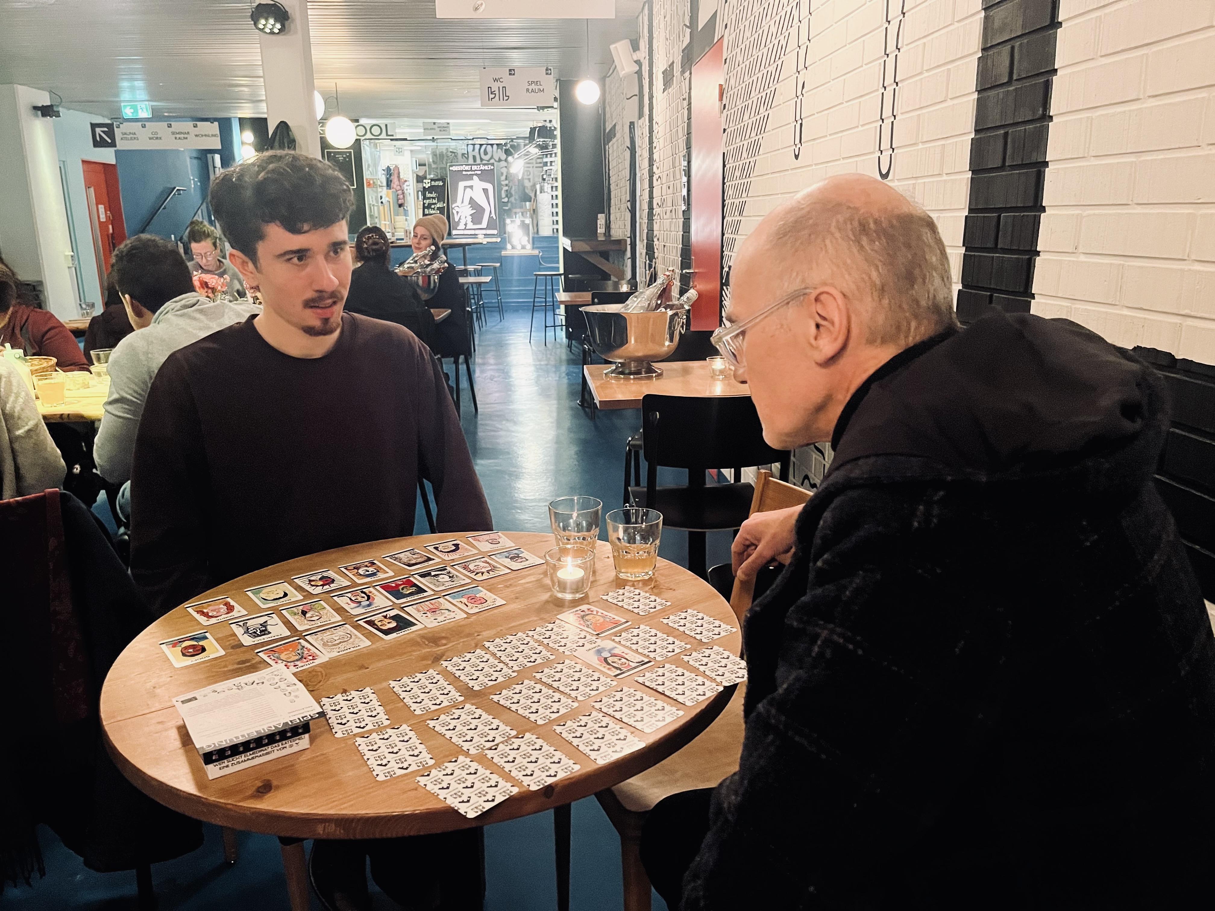 Auf dem Bild sind Pino und Helmut an einem runden Tisch zu sehen, wie sie das Spiel ausprobieren.