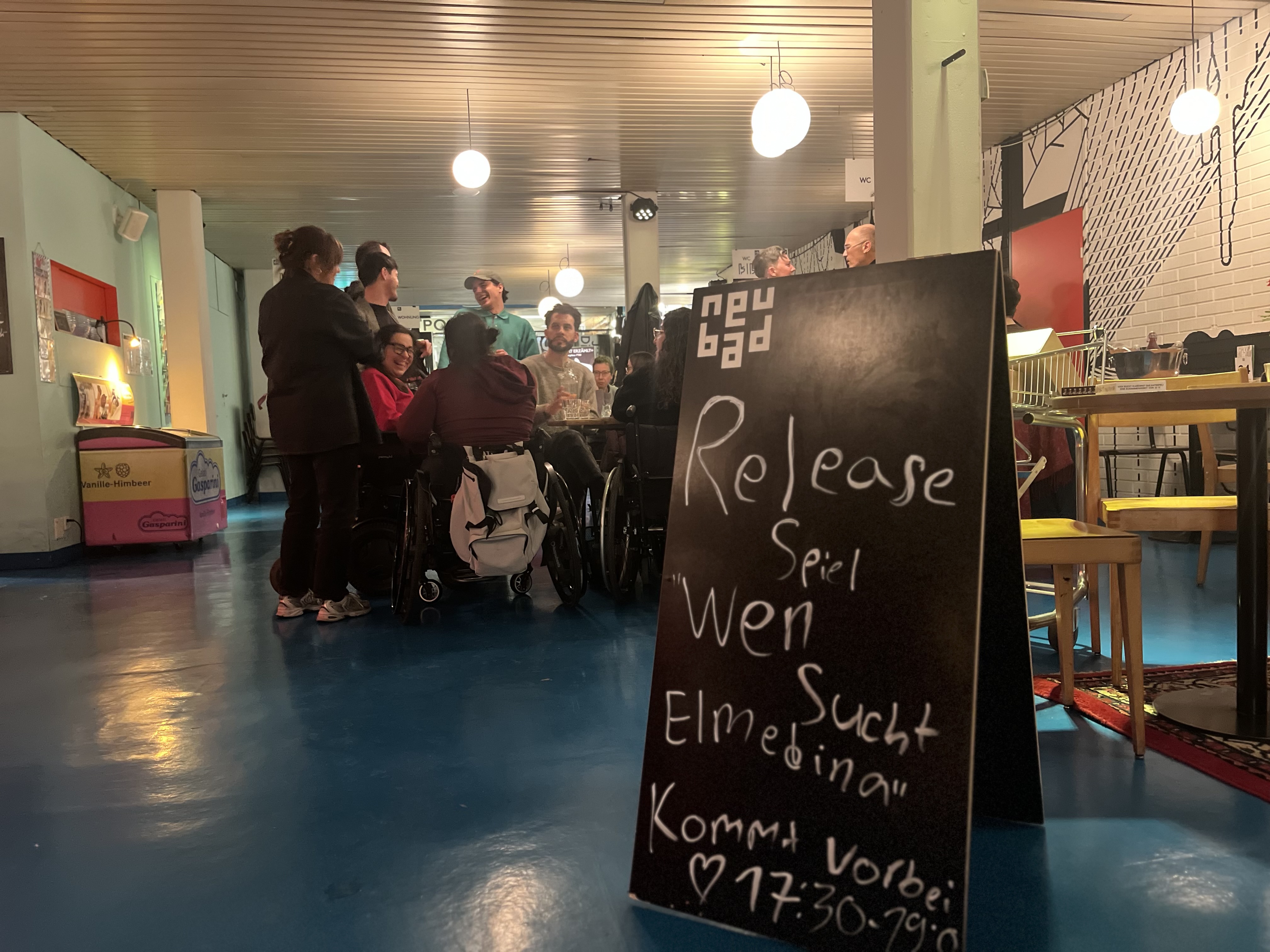Auf dem Bild sieht man eine Kreidetafel. Auf dieser Steht "Elmedina Spiel Release". Im Hintergrund sieht man Personen, die an der Release teilgenommen haben. 