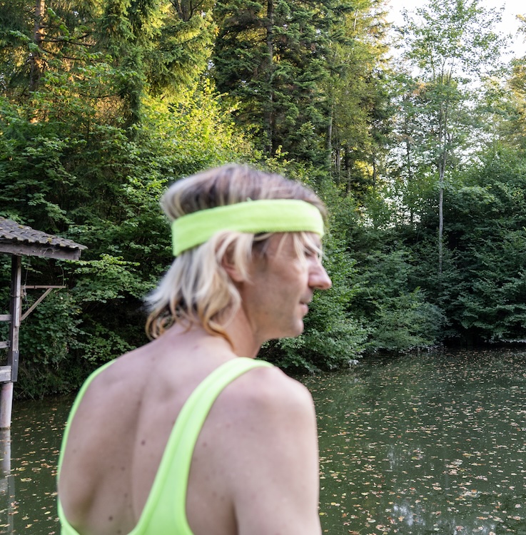 Ein Mann mit dem Rücken zur Kamera. Er trägt neongrünes Sportbody. Er hat schulterlange, blondierte Haare.