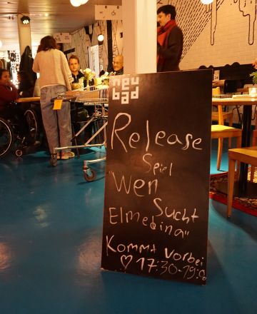 Auf dem Bild ist eine Tafel zu sehen mit der Beschriftung "Release wen sucht Elmedina". Im Hintergrund sind Personen zu sehen. 