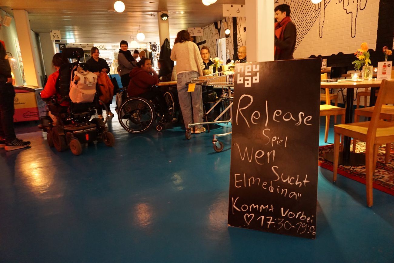 Auf dem Bild ist eine Tafel zu sehen mit der Beschriftung "Release wen sucht Elmedina". Im Hintergrund sind Personen zu sehen. 