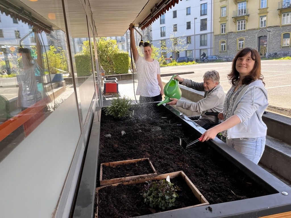 Man sieht Lukas, Marcel und mich (Nadine) am Hochbeet stehe, wie wir die Pflanzen tränken. Weil mir diese Arbeit sehr Freude macht, habe ich ein strahlendes Lächeln im Gesicht.