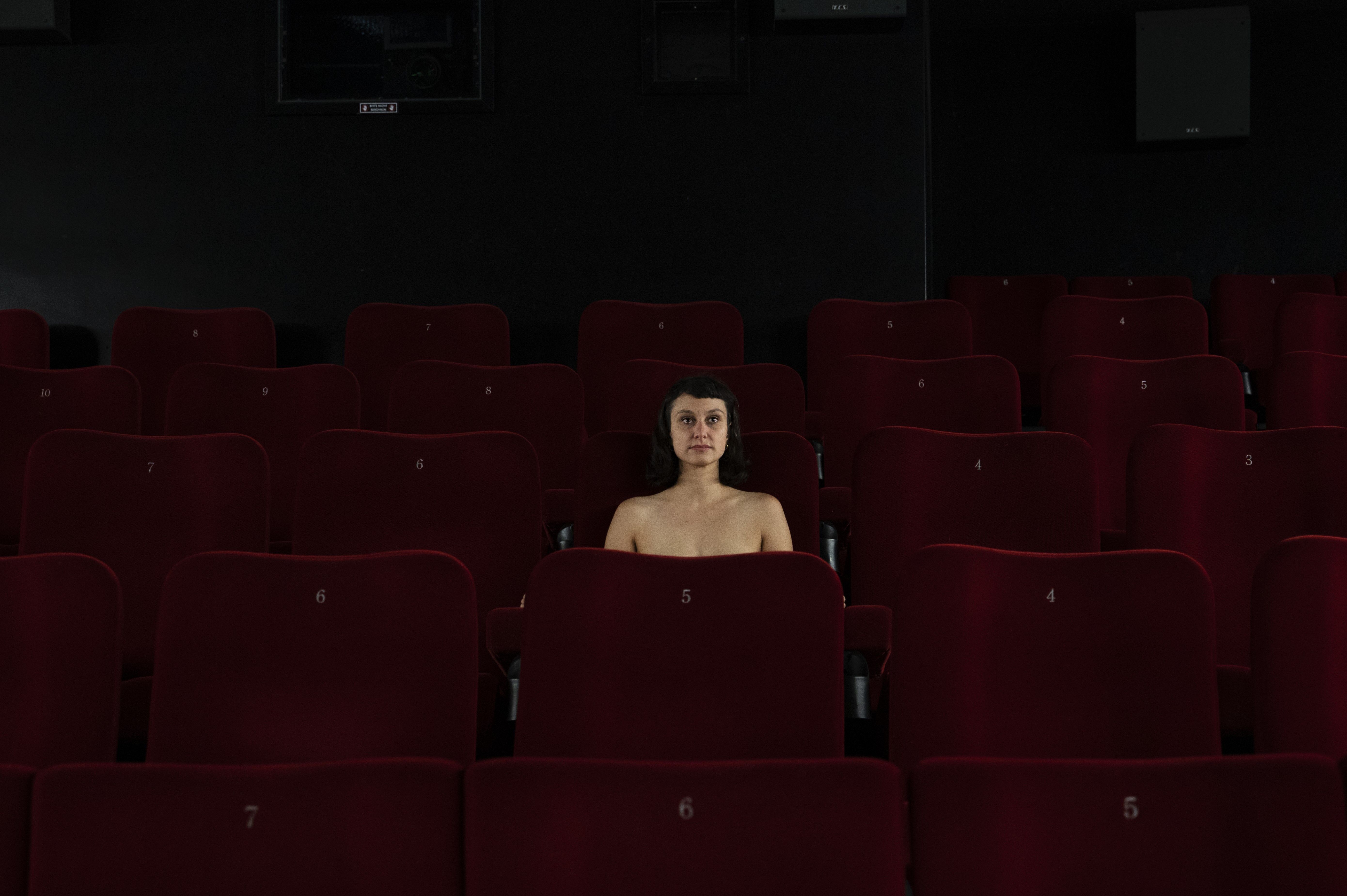 Eine Frau mit dunklen Haaren sitzt im Kino. Die Stuhlreihen um sie herum sind leer. Ihr Oberkörper ist nackt.