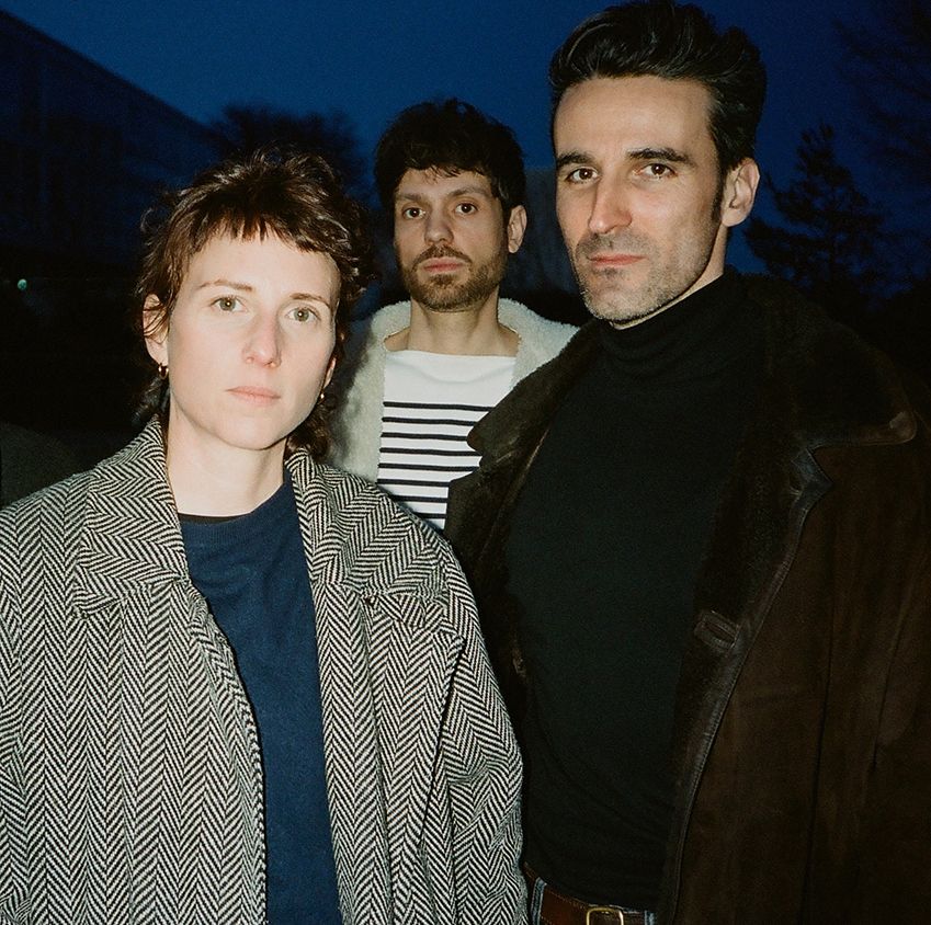 Ein Foto von drei Personen, eine Frau mit grauem Mantel, ein Mann mit schwarzem Mantel und Rollkragenpullover und ein Mann in der Mitte mit gestreiftem Oberteil.