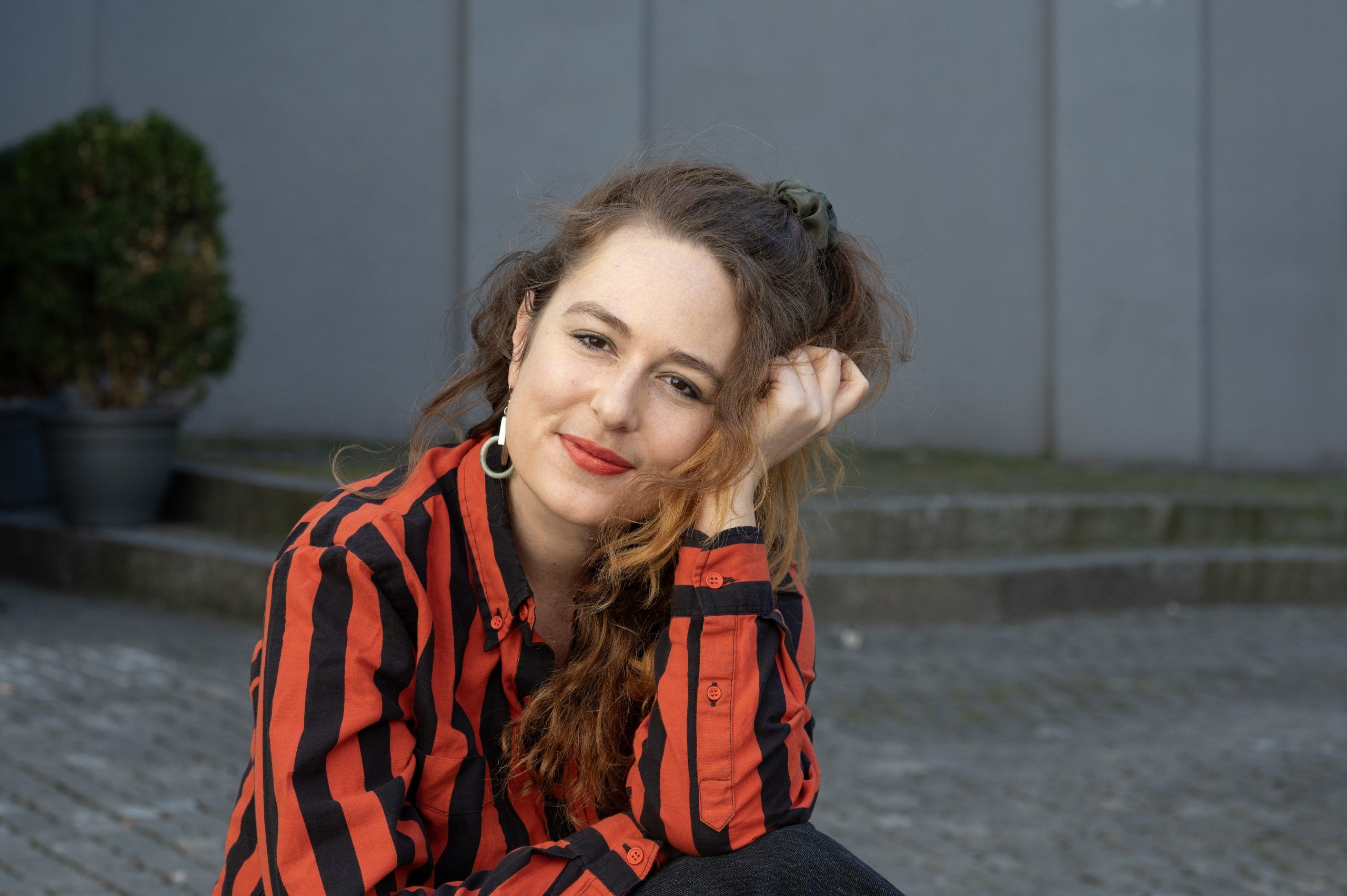 Fotografie von einer Frau mit langen braunen Haaren, goldenen Ohringen und einem schwarzrot gestreiftem Hemd. Sie stützt ihren Kopf mit ihrer Linken Hand ab.