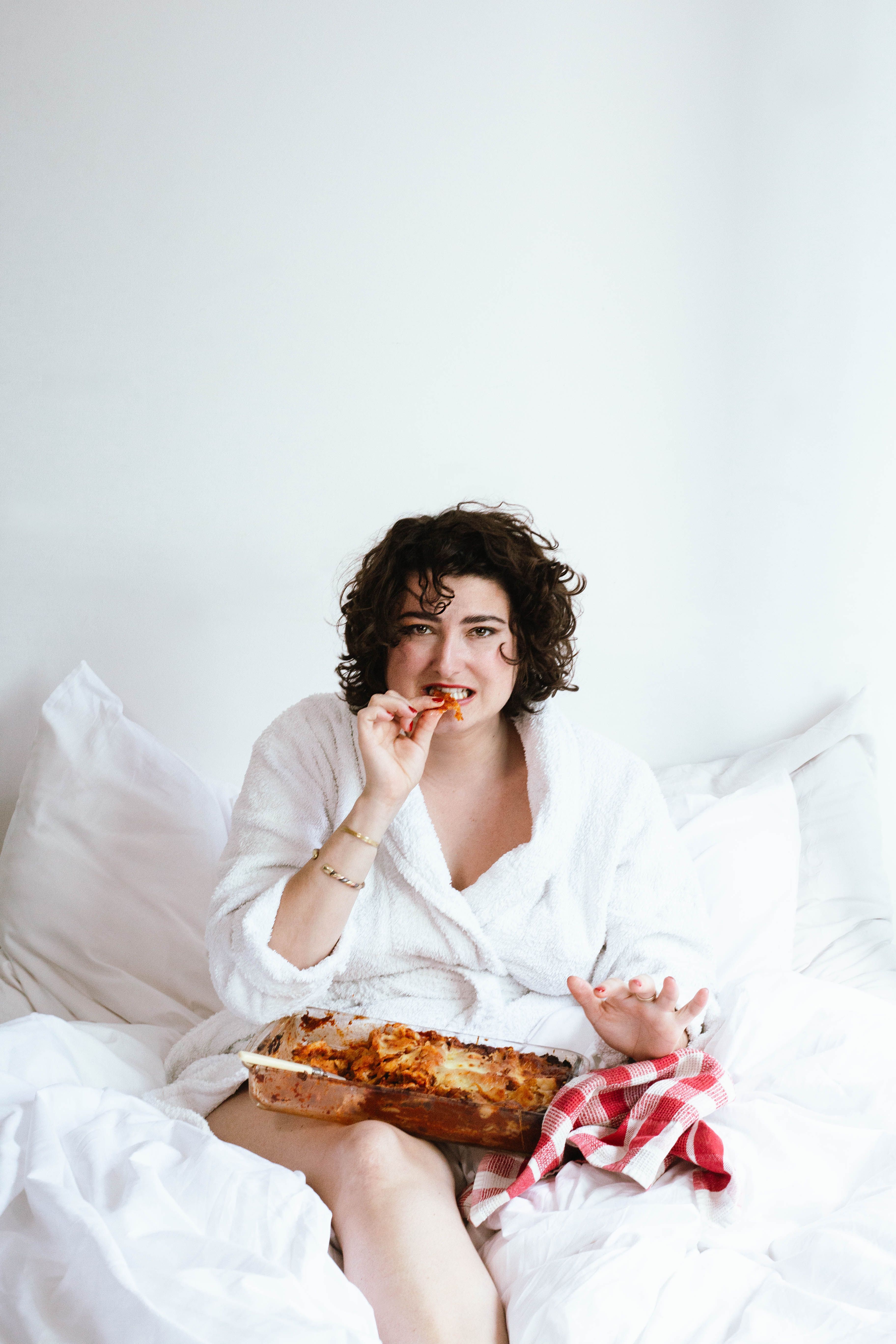 Frau mit schwarzen Locken sitzt im Bett und isst eine Lasagne aus der Auflaufform. Ein rotweiss-kariertes Küchentuch ist rechts neben der Auflaufform, die in ihrem Schoss liegt.