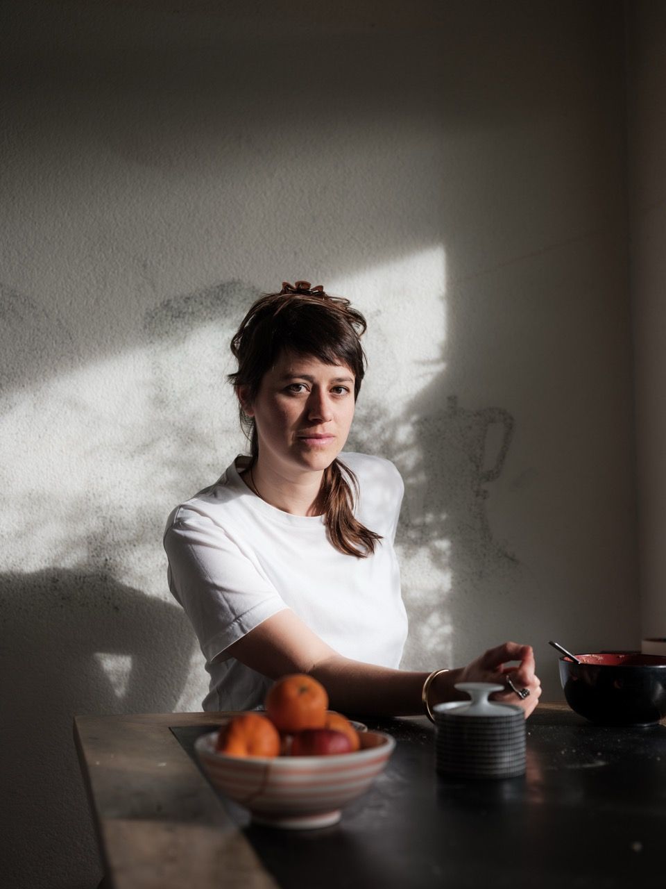 Eine Frau mit weissem T-shirt an einem Tisch vor einer Schale Orangen vor einer weissen Wand. 